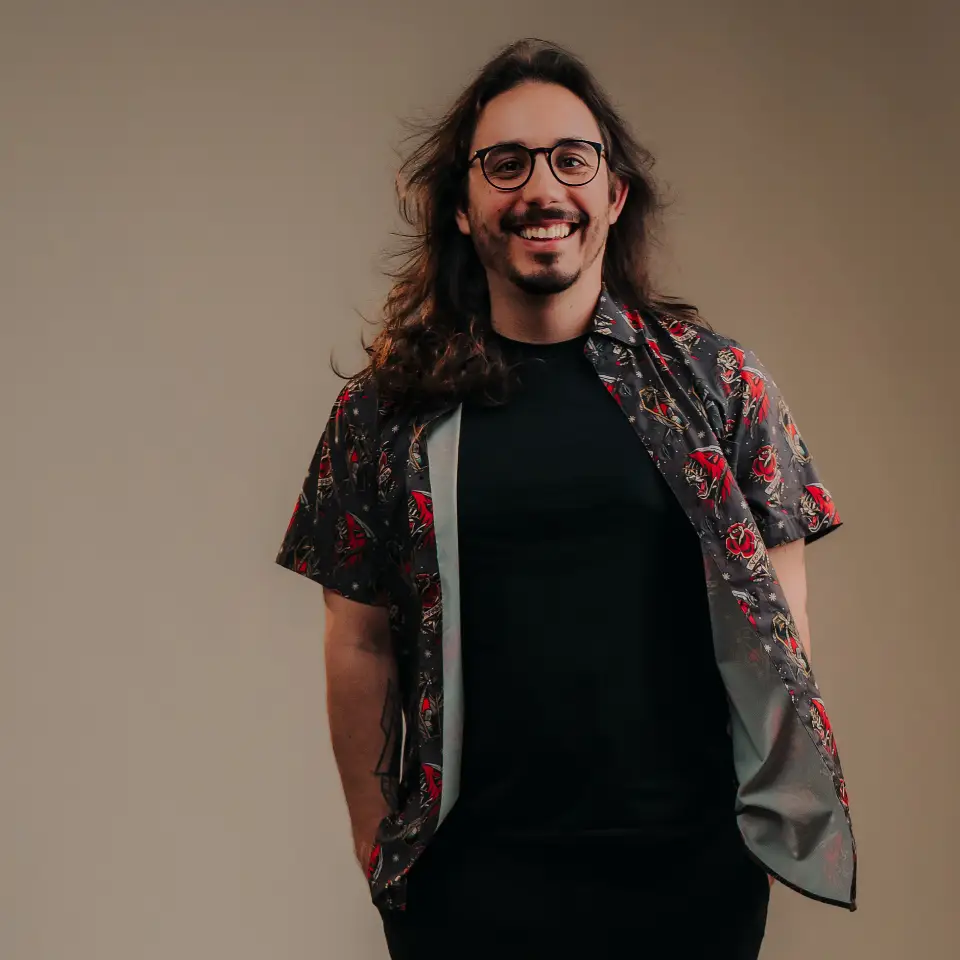 Retrato de João Dias sentado numa poltrona de couro castanha. Tem cabelo comprido e ondulado, óculos, bigode e tatuagens visíveis nos antebraços, e veste um polo preto de manga curta.
